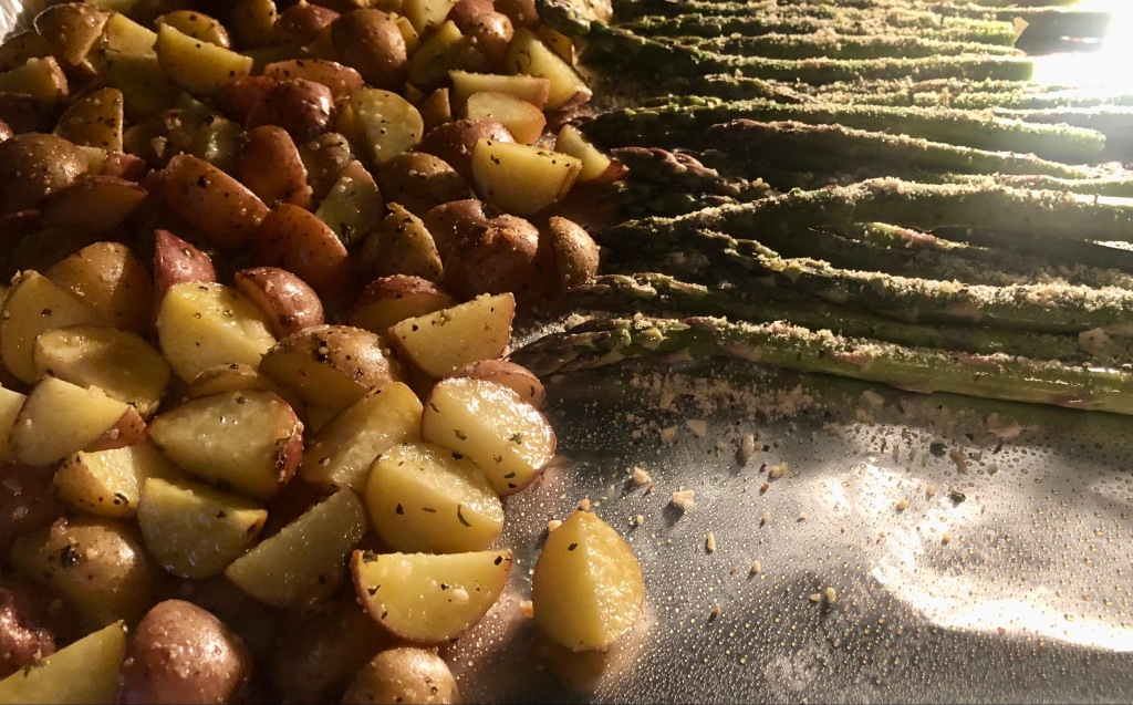 Partially roasted baby potatoes with herb and parmesan cheese mix and asparagus spears on foil-covered sheet pan.